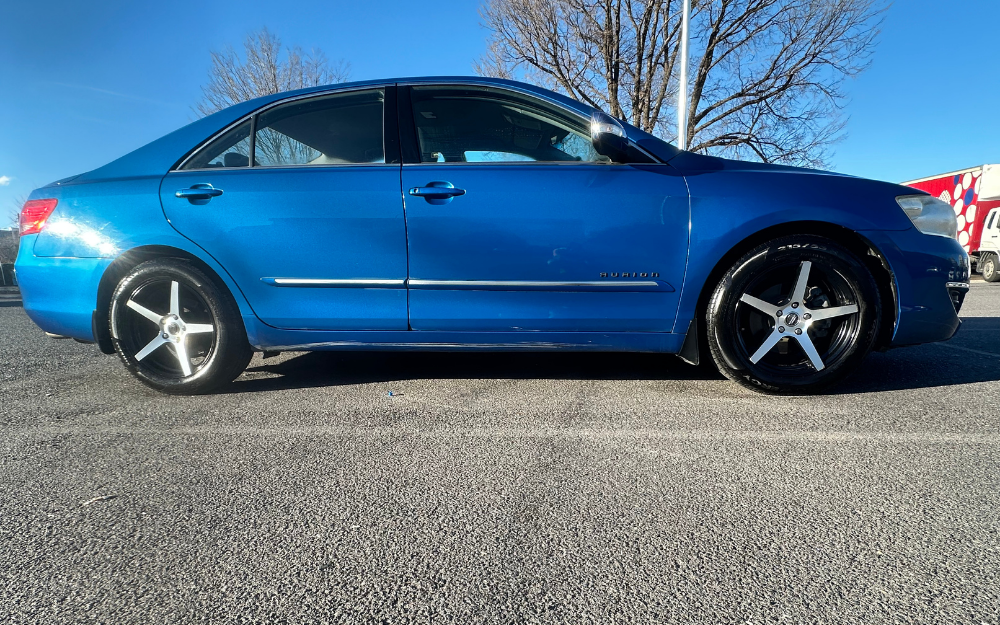 A blue Toyota Camry Hybrid sedan with black tinted windows and silver rims is parked in a lot on a sunny day, with bare trees and a building visible in the background.