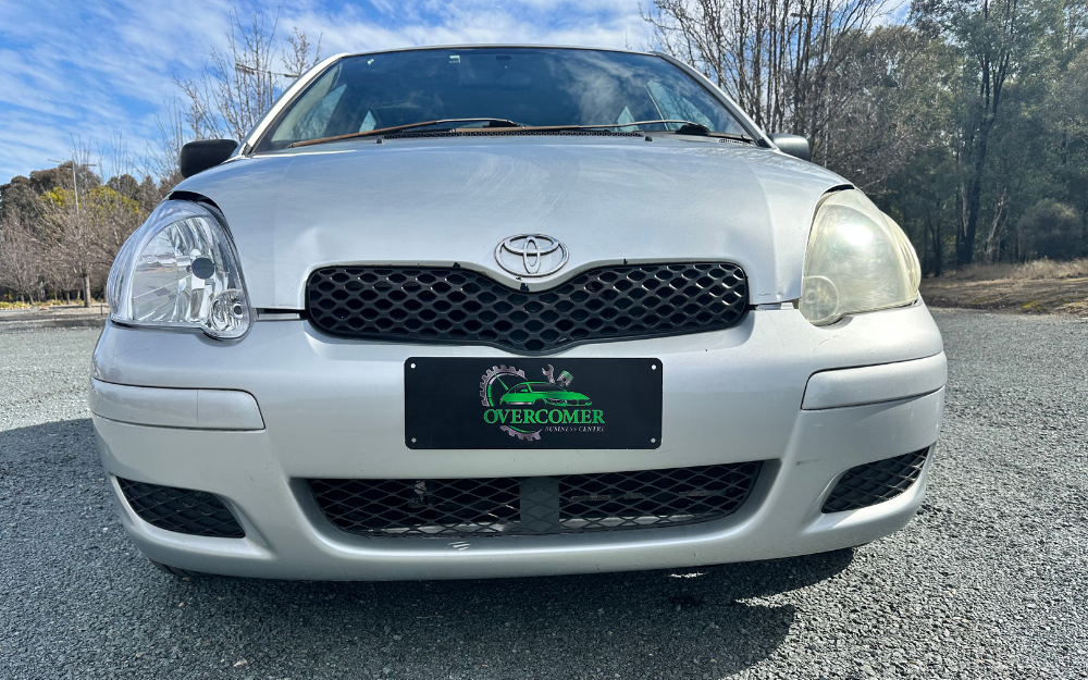 A silver Toyota car with one broken and one foggy headlight is parked on a gravel surface outdoors. The license plate reads “Overcomer Christian Center.” Trees and blue sky are visible in the background.