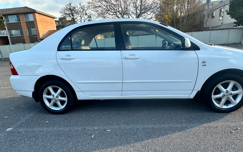 A white four-door sedan parked in an outdoor lot next to a fence, with trees and buildings visible in the background.