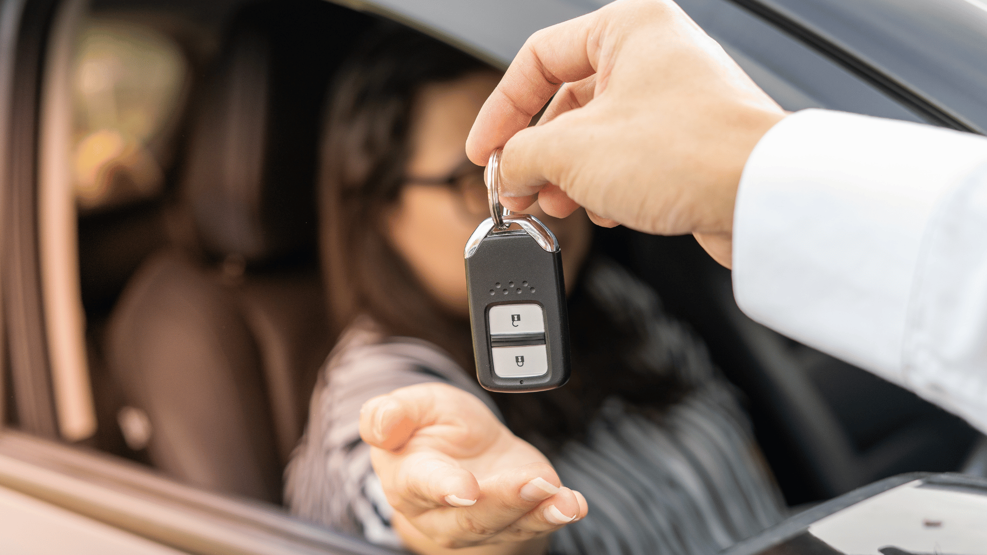 A person in a car reaches out to receive a set of car keys from another person standing outside the vehicle. The focus is on the hand holding the keys and the hand receiving them.
