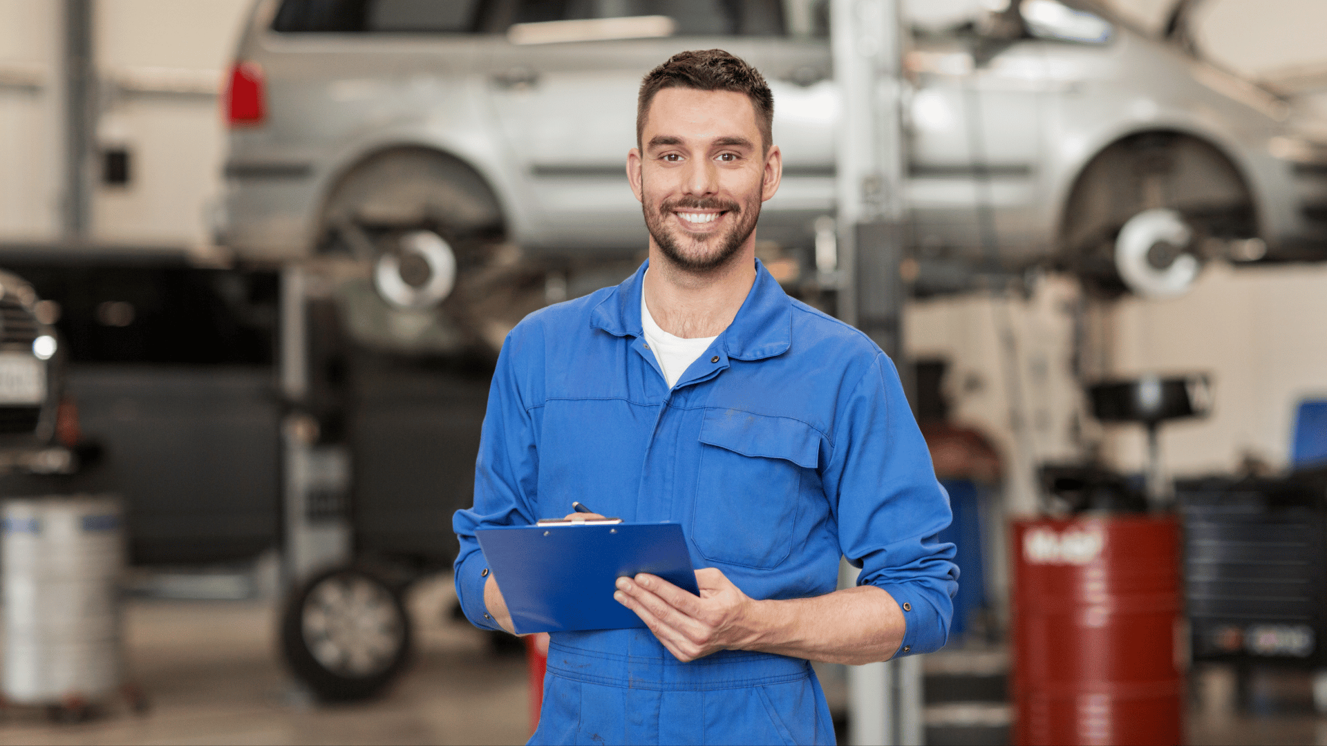 A smiling mechanic in a blue jumpsuit holds a clipboard in an auto repair shop, with a car lifted on a hydraulic lift and various tools and equipment in the background.