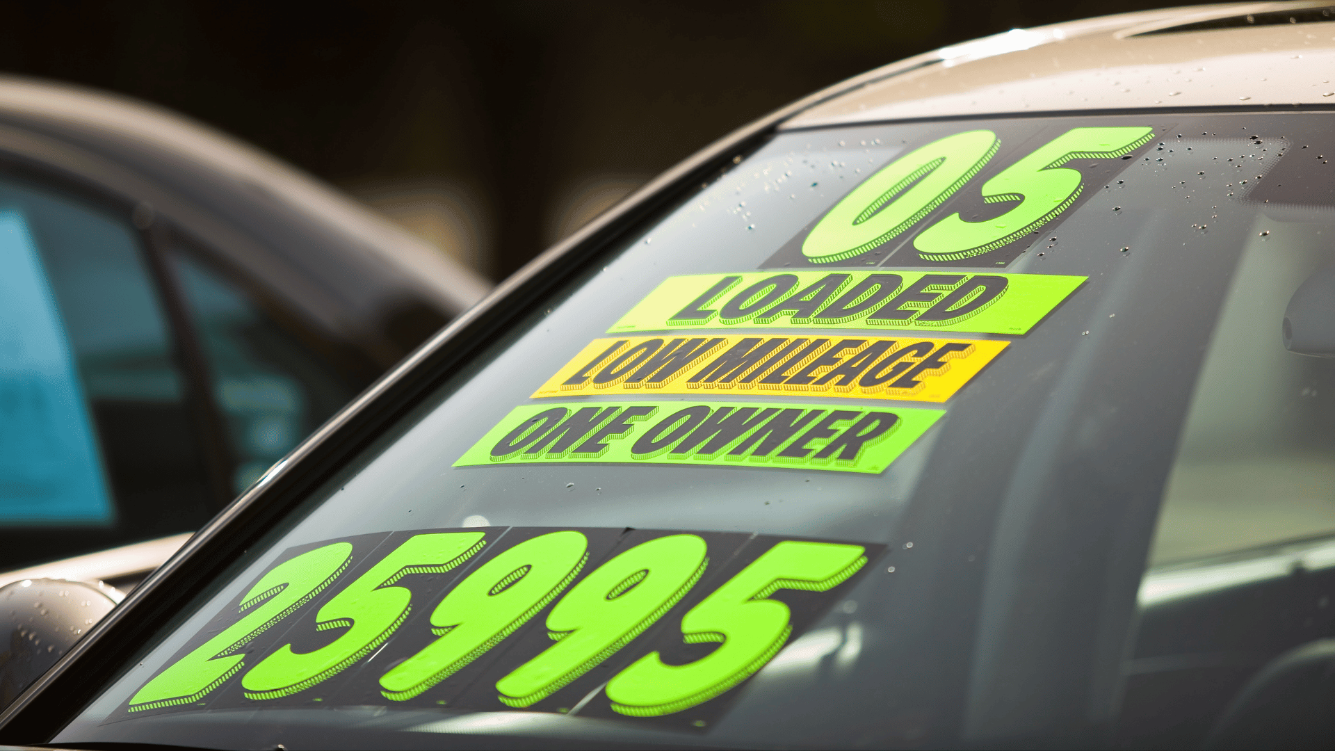 Windshield of a car displaying bright signs with text: "05," "LOADED," "LOW MILEAGE," "ONE OWNER," and "25995," indicating a 2005 model for sale with highlighted features and a price of $25,995.