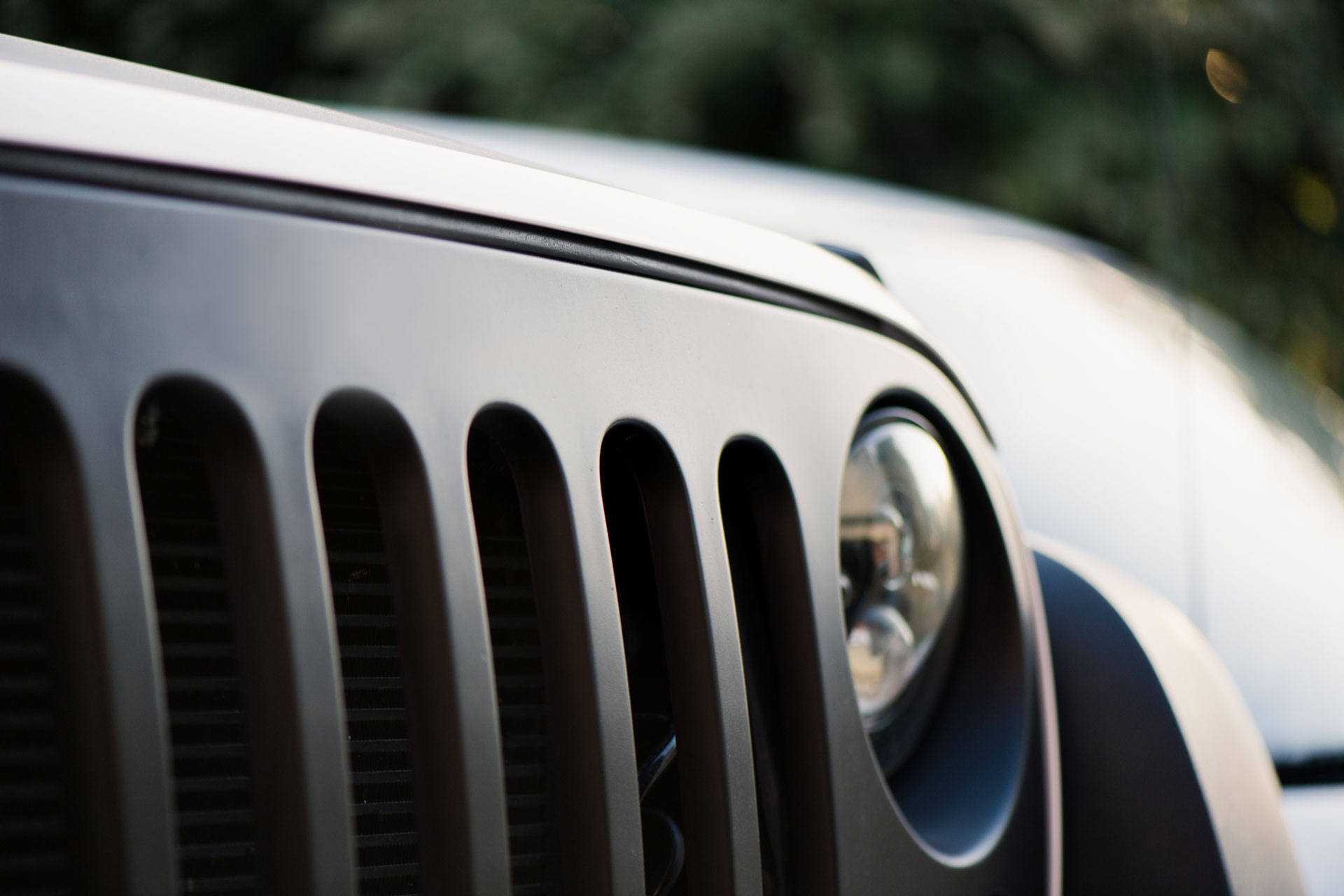 Close-up of the front grille and round headlight of a black Jeep vehicle, with a blurred background of greenery and another car.